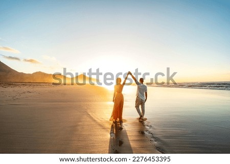 Lovely, romantic couple walking on sunset beach, enjoying evening light, relaxing on tropical summer vacation. Honeymoon. Love. Back view. Woman wearing orange maxi dress.
