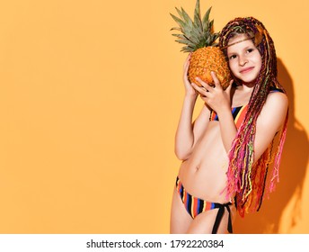 Lovely schoolgirl with cute african braids posing in striped bikini holding big pineapple close to cheek. Cropped shot isolated on orange, copy space. Children, happiness, facial expression, holidays