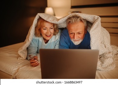 Lovely senior couple using laptop while lying on bed under blanket at night time. - Powered by Shutterstock