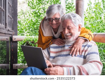 Lovely senior white haired couple hugging on a wooden balcony of mountain chalet while man using laptop computer. - Powered by Shutterstock