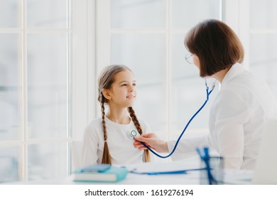 Lovely Small Girl Listens Consult Of Professional Experienced Doctor Who Listens Her Lungs With Stethoscope, Comes On Medical Checkup Appointment. Children Healthcare And Clinic Visit Concept