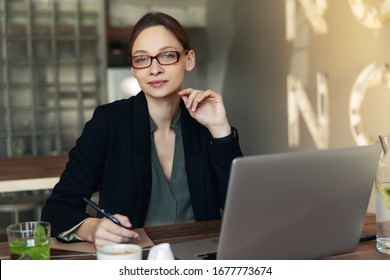Lieblich lächelnde Geschäftsfrau in stilvollen Gläsern sitzend am Café-Tisch drinnen, am Laptop arbeiten, Kamera ansehen