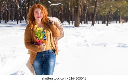 Lovely smiling woman holding her coat over shoulder while having a stroll through snowy forest with footsteps on snow surface and trees on the background
