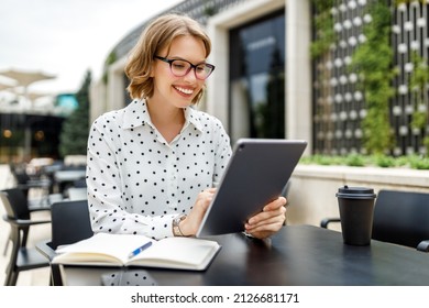 Lovely Student Girl In Glasses Sitting With Digital Tablet  And Cup Of Takeaway Coffee During Language Class While Sits In Cafe On Terrace Outside. Remote Job Concept