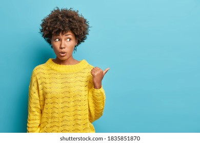 Lovely surprised Afro American woman shocked to hear unexpected relevation points thumb away and wears casual jumper impressed by incredible thing poses against blue background. Wow looks there - Shutterstock ID 1835851870