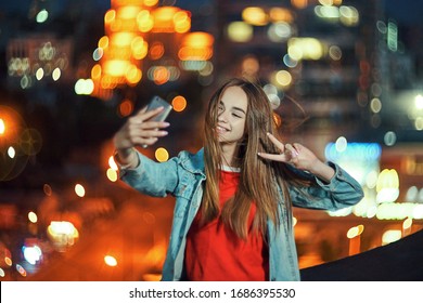 Lovely teen girl on cityscape background making self portrait with her smart phone, giving V hand sign, evening street light bokeh on background