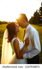 Lovely Wedding Couple At Sunset. Bride And Groom In Wedding Attire With A Bouquet Of Flowers Is In The Hands Against The Backdrop Of The 
Green Field At Sunset. Romantic Married Young Family. 