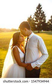 Lovely Wedding Couple At Sunset. Bride And Groom In Wedding Attire With A Bouquet Of Flowers Is In The Hands Against The Backdrop Of The 
Green Field At Sunset. Romantic Married Young Family. 