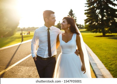 Lovely Wedding Couple At Sunset. Bride And Groom In Wedding Attire With A Bouquet Of Flowers Is In The Hands Against The Backdrop Of The 
Green Field At Sunset. Romantic Married Young Family. 