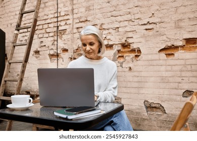 A lovely woman with gray hair sits at a table, immersed in her laptop, sipping coffee. - Powered by Shutterstock