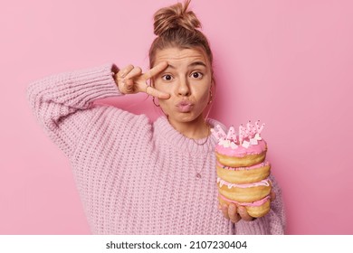 Lovely woman with hair gathered in bun keeps lips rounded makes peace gesture over eye holds birthday doughnuts celebrates special occasion wears knitted sweater isolated over pink background