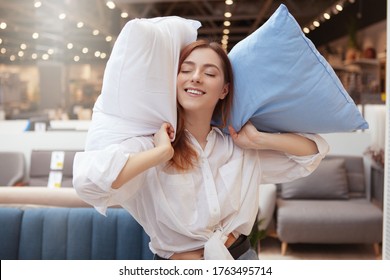 Lovely woman smiling joyfully while shopping for bedding at furniture store. Woman holding pillows, smiling with eyes shut