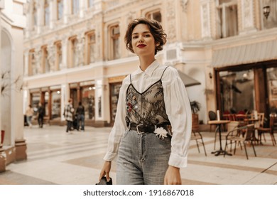 Lovely woman with wavy hair in jeans with belt and flower smiling at street. Cool lady in white blouse with lace posing in city..