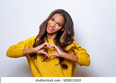 Lovely young african american woman with happy smile, shapes heart with both hands, expresses love to you, wears yellow wool sweater, says be my valentine flirts with boyfriend poses indoor copy space