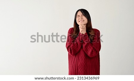 A lovely young Asian woman in a red sweater is holding clasped hands over her chest, smiling, and closing her eyes in a shy and tender pose. isolated on white background