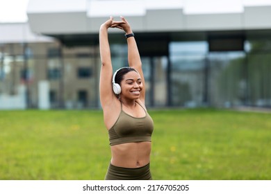 Lovely Young Black Woman Listening To Music In Headphones, Stretching Arms Above Her Head, Doing Sports At City Park, Free Space. Athletic African American Female Enjoying Outdoor Training