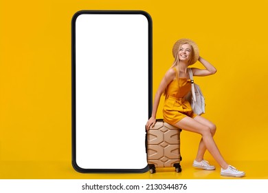 Lovely Young Blonde Woman Sitting On Suitcase Near Giant Smartphone With Mockup, Booking Summer Vacation Online, Using Internet Travel Agency On Orange Studio Background