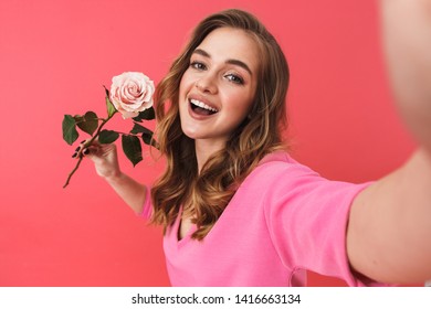 Lovely young blonde woman standing isolated over pink background, taking a selfie, showing rose