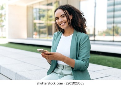 Lovely young brazilian or latino business woman, wearing elegant suit, holding cell phone sitting outdoors, using gadget for online messaging, checking email, browsing websites, look at camera, smiles - Powered by Shutterstock