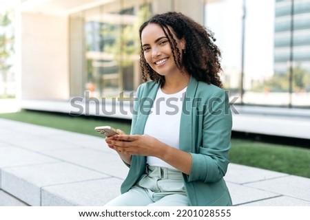 Lovely young brazilian or latino business woman, wearing elegant suit, holding cell phone sitting outdoors, using gadget for online messaging, checking email, browsing websites, look at camera, smiles