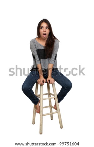 A lovely young brunette in casual wardrobe, with a surprised or shocked facial expression, sits on a barstool, isolated on a white background with generous copyspace.
