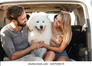 Lovely young happy couple sitting in the back of their car at the beach, playing with dog