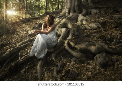 Una joven y encantadora dama con un elegante vestido blanco disfrutando de las vigas de luz celestial en su cara sentada en las poderosas raíces de un árbol antiguo en bosques encantados