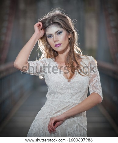 Lovely young lady wearing elegant white dress enjoying the beams of celestial light and snowflakes falling on her face. Pretty brunette girl in long wedding dress posing on a bridge in winter scenery