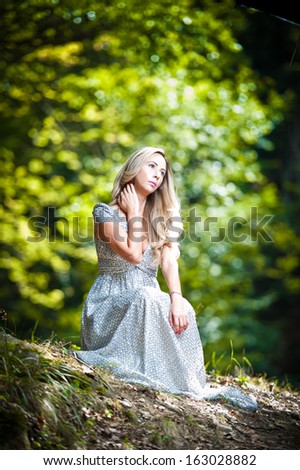 Lovely young lady wearing elegant white dress enjoying the beams of celestial light on her face in enchanted woods. Pretty blonde fairy lady with white dress. Glamorous princess in the woods