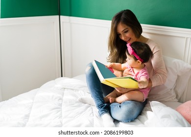 Lovely Young Mom Reading A Children's Book To Her Cute Baby Girl While Relaxing In Bed Together