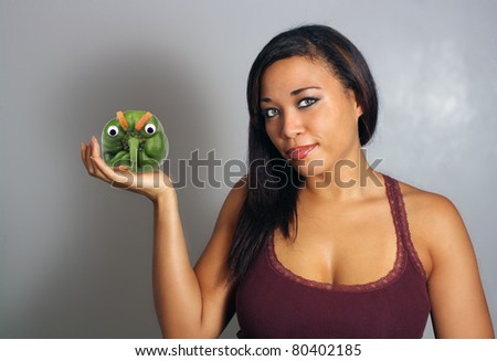 A lovely young multiracial woman holds a green pepper with doll eyes and eyebrows made of carrot slices.