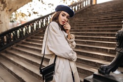 Lovely Young Parisienne With Brunette Hair In Stylish Beret, Beige Trench Coat And Black Bag, Standing On Old Stairs And Sensitively Posing Outdoors. Warm Autumn