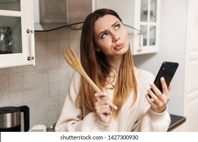 Lovely young woman cooking while standing at the kitchen at home, holding mobile phone, thinking
