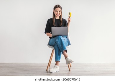 Lovely Young Woman With Credit Card And Laptop Sitting On Chair, Buying Goods On Internet, Making Contactless Payment Against White Studio Wall. Online Shopping, Remote Banking Concept