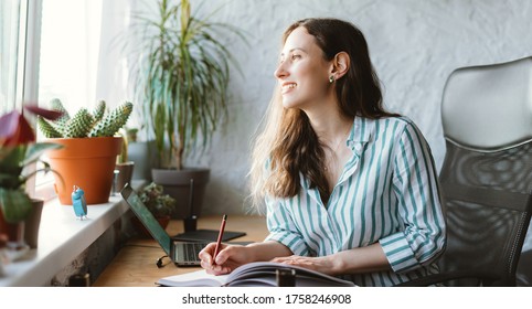 Lovely young woman is looking through window while writing in journal at her work desk.