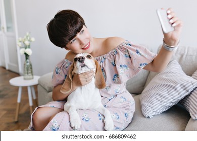 Lovely Young Woman With Nude Makeup Touching With Love Her Beagle Dog And Taking Picture Of Herself. Indoor Portrait Of Smiling Brunette Girl And Her Pet Playing Together In Cozy Room.