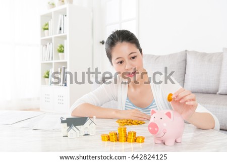 lovely young woman sitting in living room  at home and holding gold coin saving money into pink piggy bank for buying new house with family.