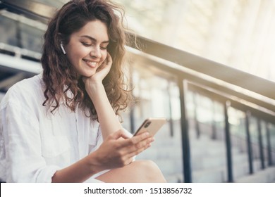 Lovely young woman sitting on steps outside. Sun shining bright. Sunset. Holding phone in hands and smile. Listening to music through wireless earphones - Powered by Shutterstock