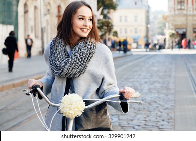 Lovely young woman, wearing in gray jacket and scarf, with black bag, posing with bike on the street of old European city, on sunny day, waist up