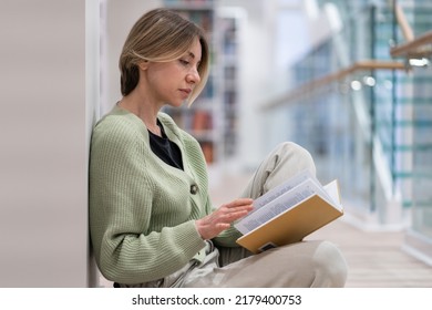 Lover of books. Focused pensive middle-aged female passionate reader sitting on floor with book in hands, mature woman bibliophile reading in public library, soft focus. Self education,