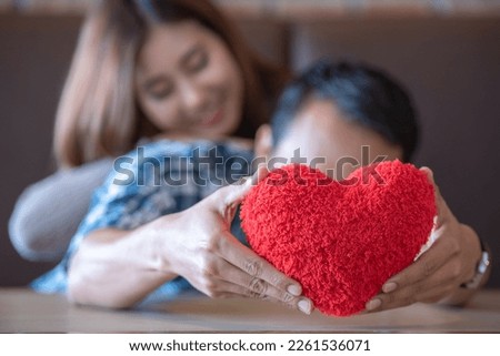 Lover couple happy in love relaxing on sofa looking in eye smiling teasing with big red heart shape pillow in valentine day honeymoon. teasing teasing with red heart pillow.