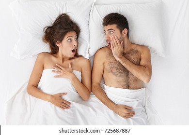 Lovers In Bed Look With Terrified Expression At Each Other As Hear That Someone Came In House, Being Scared To Be Caught. Horrified Young Married Couple Overslept Work, Didn`t Hear Alarm Clock.