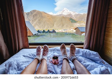 Lovers Legs In Warm Knitted Socks Lying On Soft White Bed In Wooden Cottage With Big Window And Mountains View. Good Morning Together 