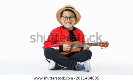 Lovey cutout portrait of young Asian healthy boy wearing brown hat, glasses, red shirt, and long black pants happily sitting with smile while enjoy performing as musician by playing small guitar