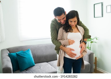 Loving And Adorable Couple Talking To Their Baby In The Belly. Latin Man And Caucasian Pregnant Woman In Love Hugging In The Living Room