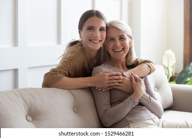 Loving adult 30s daughter hug elderly mother from behind while mom sitting on couch people posing looking at camera smiling feels happy, concept of multi generational family, relative devoted person