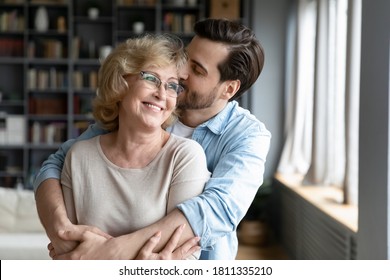 Loving Adult Son Kissing And Hugging Happy Mature Mother From Back Close Up, Expressing Gratitude, Middle Aged Woman Wearing Glasses And Young Man Enjoying Tender Moment, Standing In Living Room