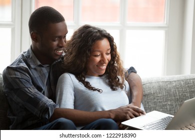 Loving African American couple using laptop together, sitting on couch at home, handsome man embracing attractive woman, watching video, cinema, shopping online, looking at screen with smile
