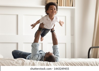 Loving african American dad lying on bed playing with cute little toddler son acting plane, happy black father have fun engaged in funny activity with small kid hold lift in arms in bedroom