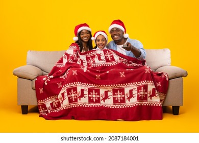 Loving African American Family In Santa Claus Hats Watching Christmas Movies Celebrating Xmas Eve At Home, Sitting On Couch Covered With Red Blanket, Holding Using Remote Control, Yellow Studio Wall - Powered by Shutterstock